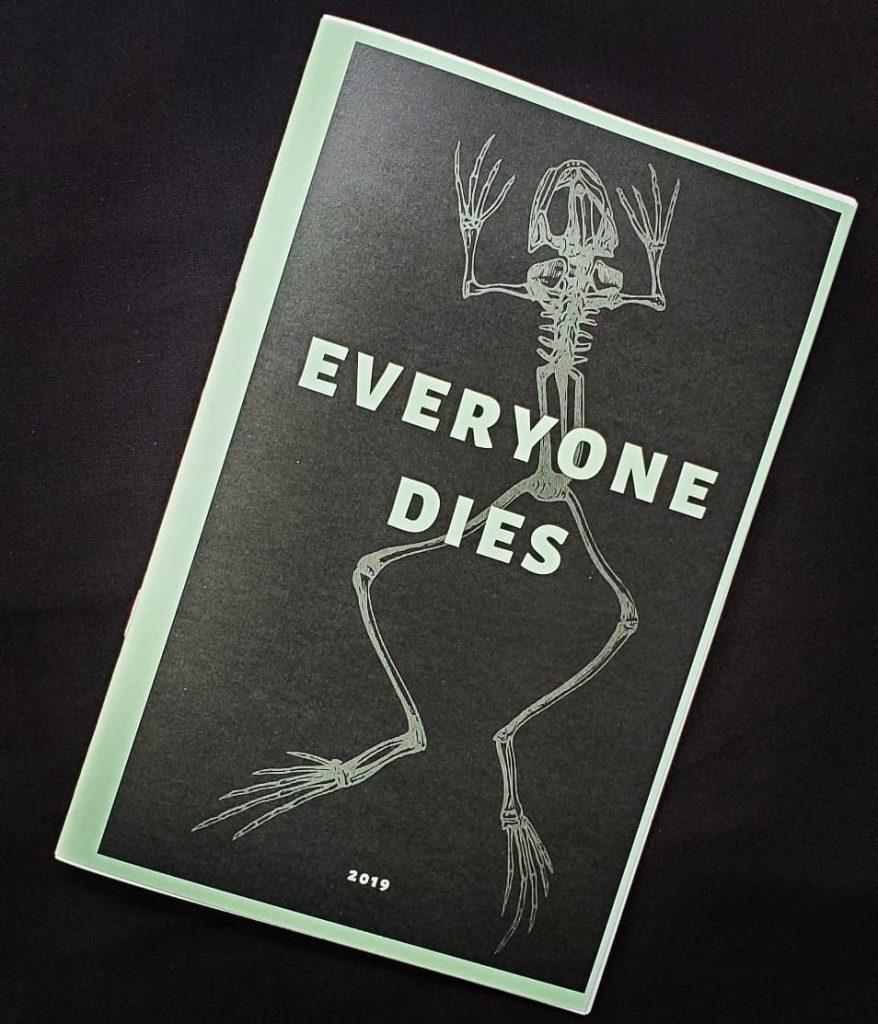 zine cover is at an angle on a black background, showing a black background with a frog skeleton on the cover with white text that reads "everyone dies"